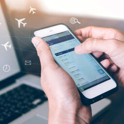 person booking cheap flight to Amsterdam on cell phone with laptop in background