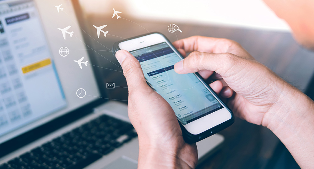 person booking cheap flight to Amsterdam on cell phone with laptop in background
